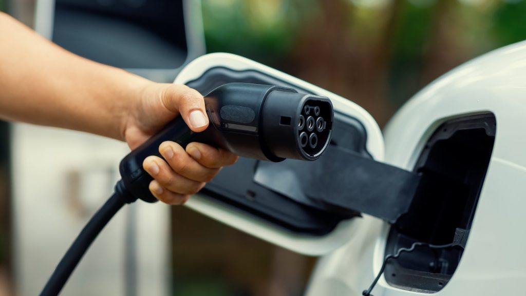 Focus hand insert progressive EV charger plug to EV car with blur background. Hand inserting EV charging plug to electric vehicle in focus shot with blurred background of outdoor natural greenery. Progressive sustainable energy powered electric charging station for rechargeable